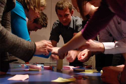 A group crowds around a table, constructing a tower of post-it notes and paperclips.