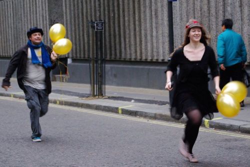 Two players with balloons run along a Soho street.